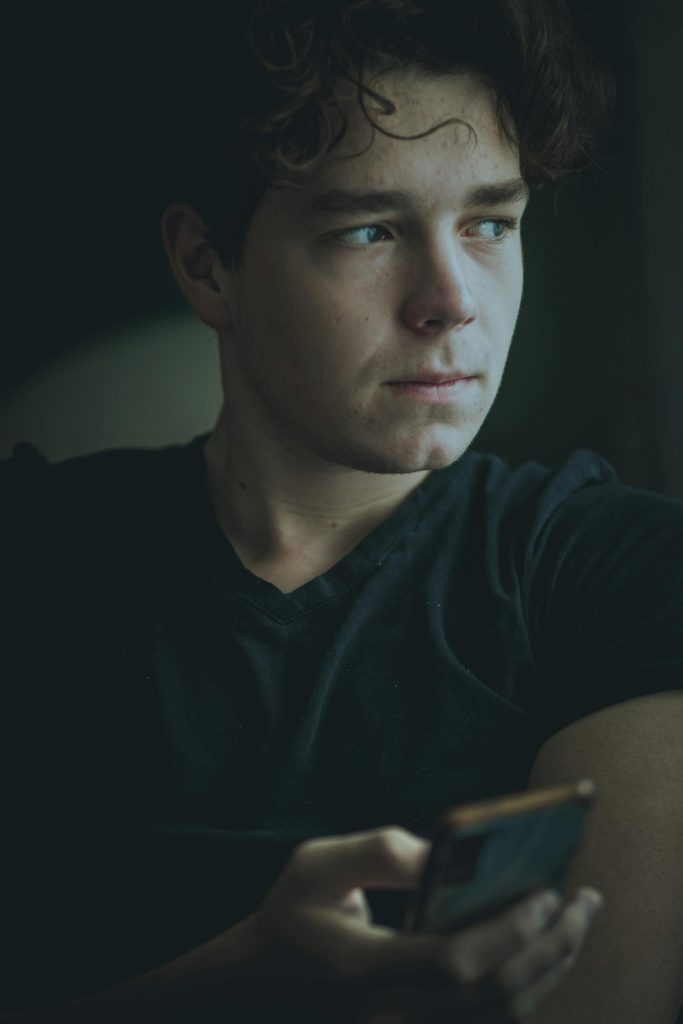 pexels photo 13587120 13587120 Young man with curly hair holding a phone, looking contemplative indoors in low light.